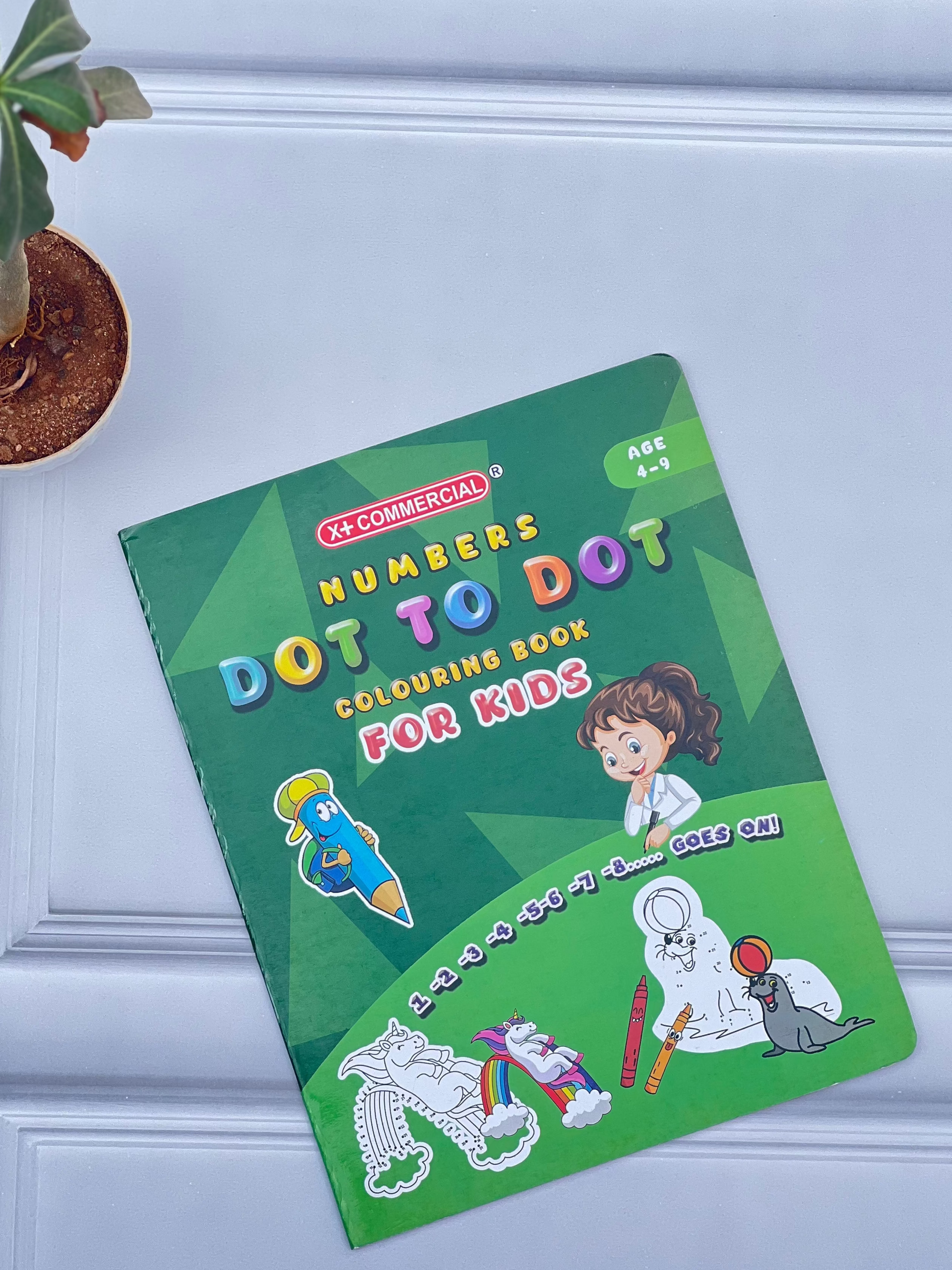 Children's dot-to-dot coloring book on a light blue surface with a plant in the background